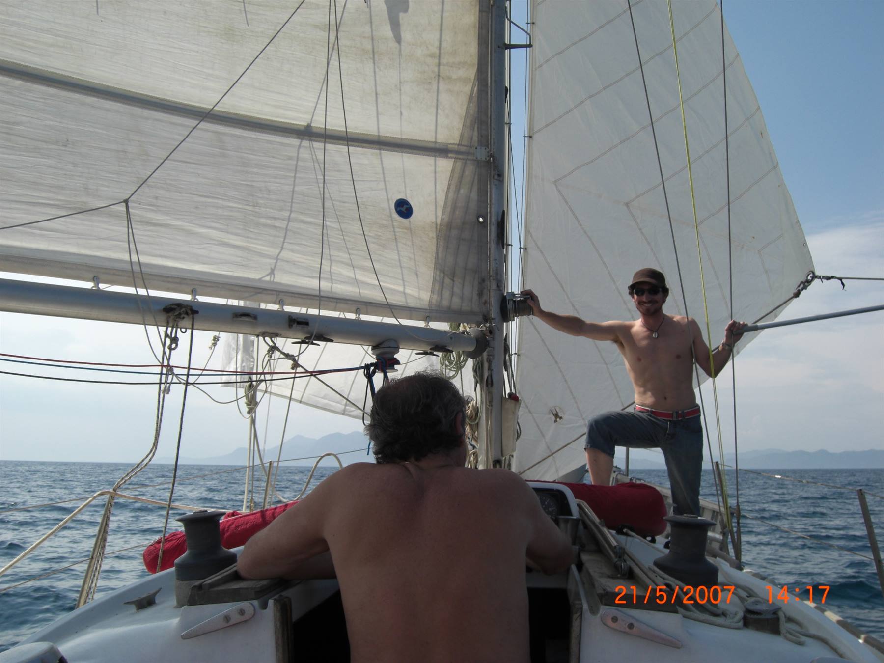 Two men aboard a sailboat, one handling rigging lines at open sea