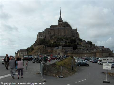 Tourists walk toward Mont-Saint-Michel abbey under a cloudy sky