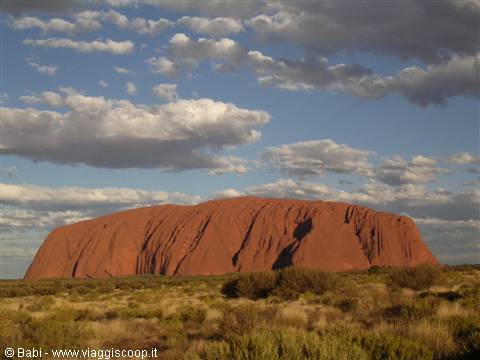 Red rock monolith of Uluru under a cloudy sky, surrounded by desert shrubs