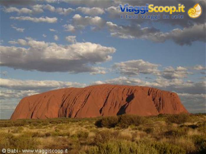 Red rock monolith of Uluru under a cloudy sky, surrounded by desert shrubs