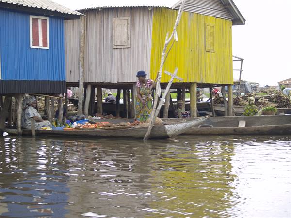 Woman in a canoe loaded with fruit beside wooden stilt houses