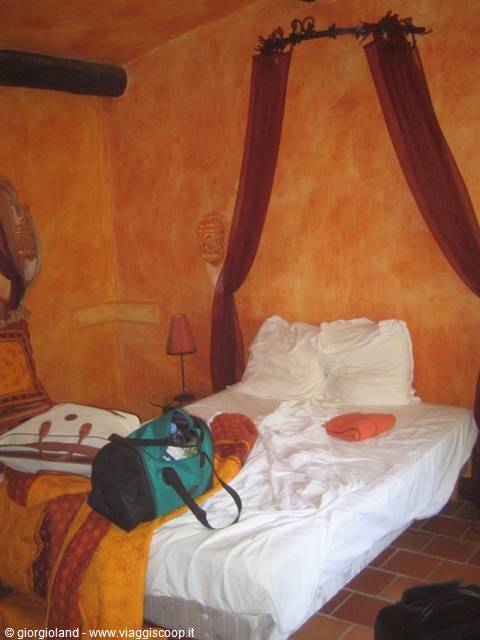 Room with ochre walls, unmade white bed with burgundy drapes, green duffel bag