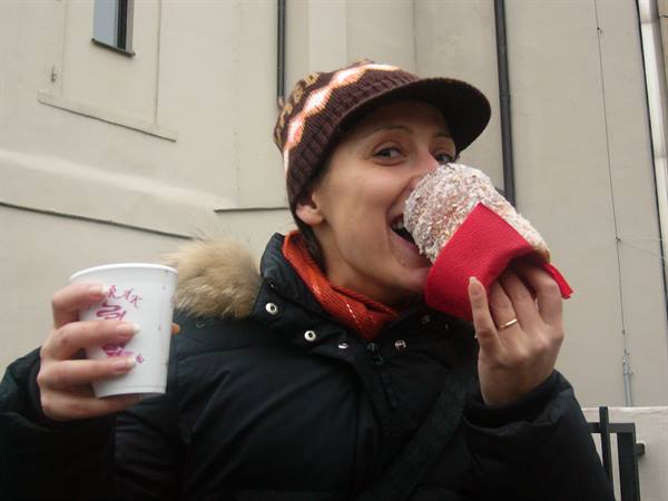 Woman in brown hat bites a sugary pastry wrapped in red, holding a paper cup