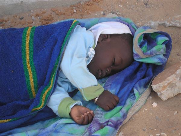 Sleeping child wrapped in blue and green blankets on sandy ground
