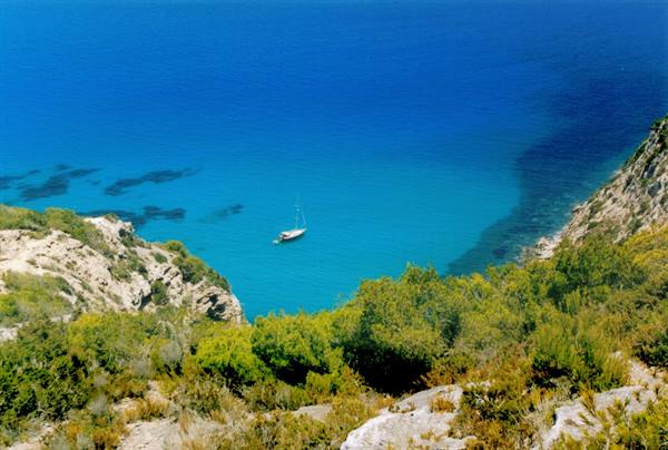Sailboat anchored in a turquoise cove between rocks and green shrubs