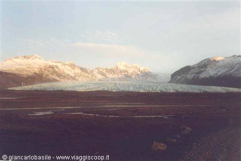 Blue glacier tongue between snowy mountains, pink light, gravel plain in foreground, Iceland