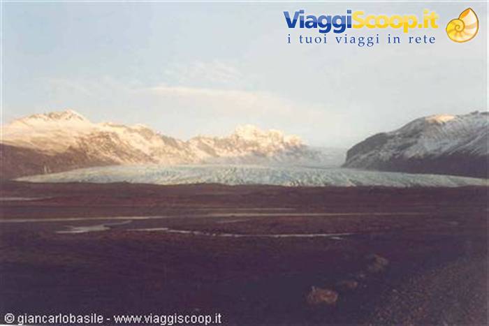 Blue glacier tongue between snowy mountains, pink light, gravel plain in foreground, Iceland