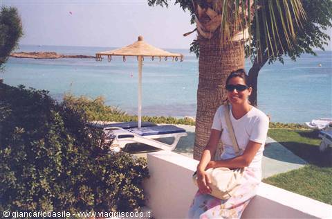 Woman in sunglasses sitting on white wall, palm tree and straw umbrella by turquoise sea