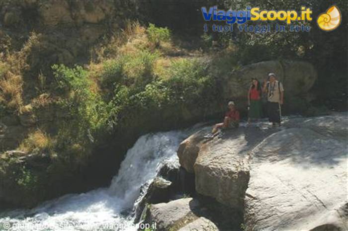 Tourists standing and sitting on a rock beside a waterfall among green bushes