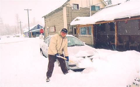 Man shoveling deep snow in front of a car and snowy wooden buildings