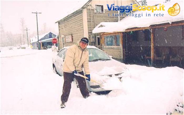 Man shoveling deep snow in front of a car and snowy wooden buildings
