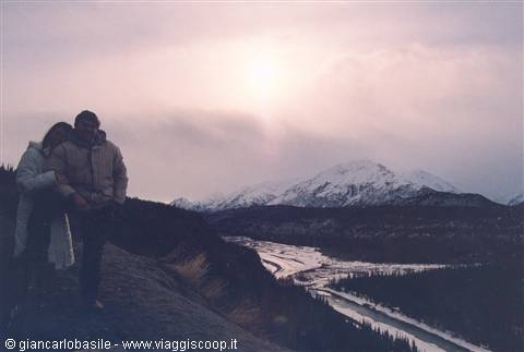 Couple embracing in the snow, with snowy mountains and a frozen river behind