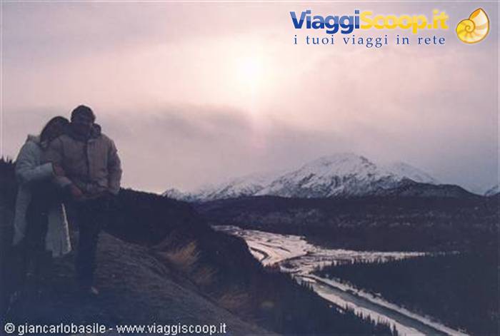 Couple embracing in the snow, with snowy mountains and a frozen river behind