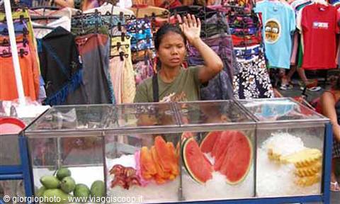 Woman waving behind fruit stall with watermelon, pineapple and clothes