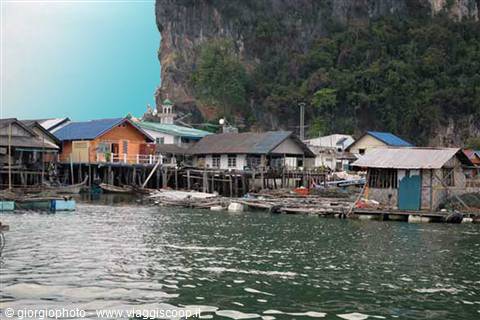 Stilt wooden fishing village over green water, vegetation-covered limestone cliff, Koh Panyi island