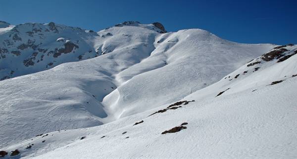 Snow-covered mountain slope under blue sky, dark rocks emerge from snow