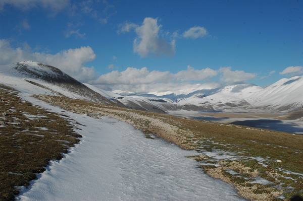 Snow patches on green meadow at Castelluccio, snowy mountains under white clouds