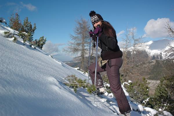 Woman in black beanie and purple pants climbs snowy slope with trekking poles, snowy peaks behind