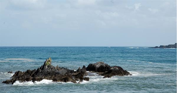Dark rocks emerge from the blue sea, waves foaming at their base