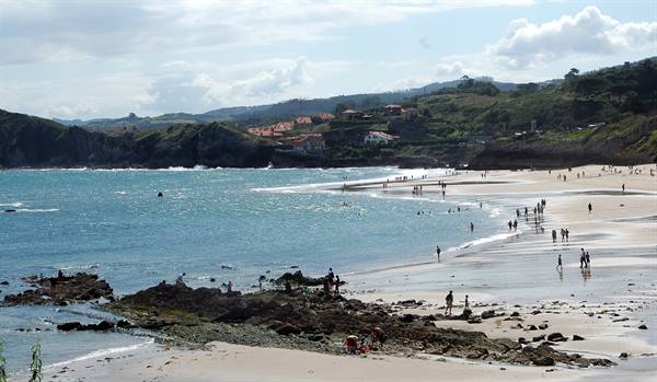 Bathers on sand and rocks by green sea, houses and hills behind