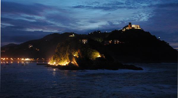 Illuminated rock in the sea at night, hotel on hill behind