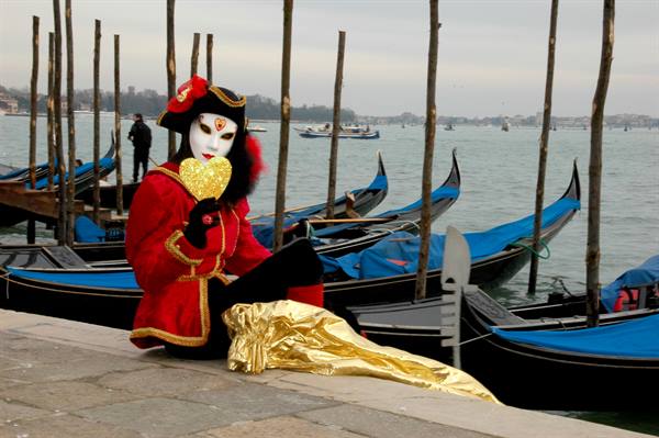 Carnival mask in red costume with white bauta beside gondolas in Venice