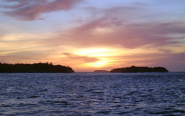 Sun sinking behind dark islets, pink clouds over rippled sea