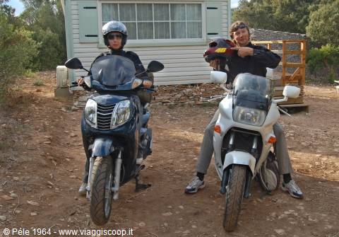 Two riders in helmets sit on a scooter and motorcycle in front of a mobile home