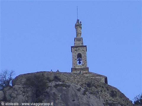 Madonna statue atop stone bell tower with bell, on rocky cliff